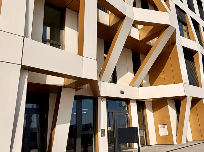 Vue détaillée de l’entrée de la Brainfactory Bochum, avec une structure de façade distinctive, des éléments de soutien inclinés et une architecture moderne dans des tons blancs et dorés.