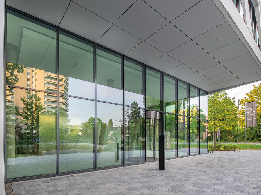 Entrée du ONE PLAZA avec une vaste façade vitrée, une architecture moderne et une vue sur les bâtiments et les espaces verts environnants.